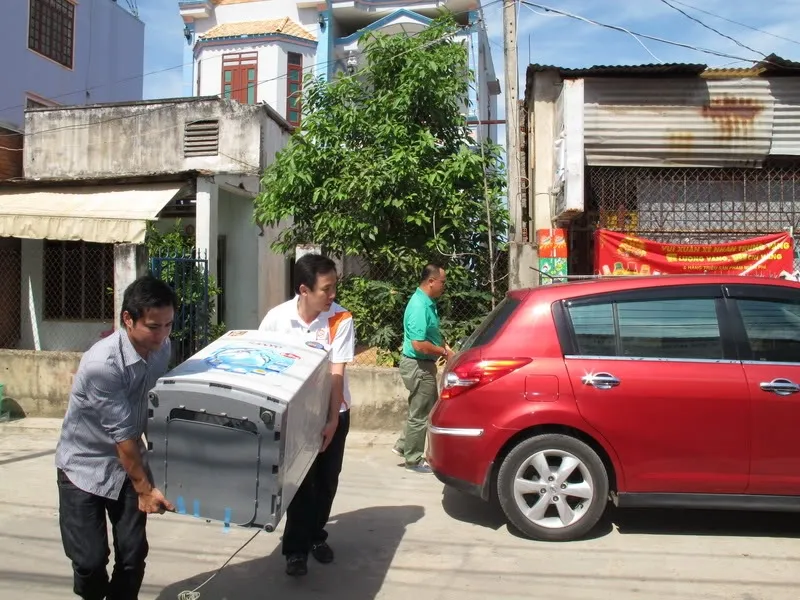 Alt text: Hai người đang cẩn thận nâng máy giặt lên xe máy và giữ ở tư thế thẳng đứng